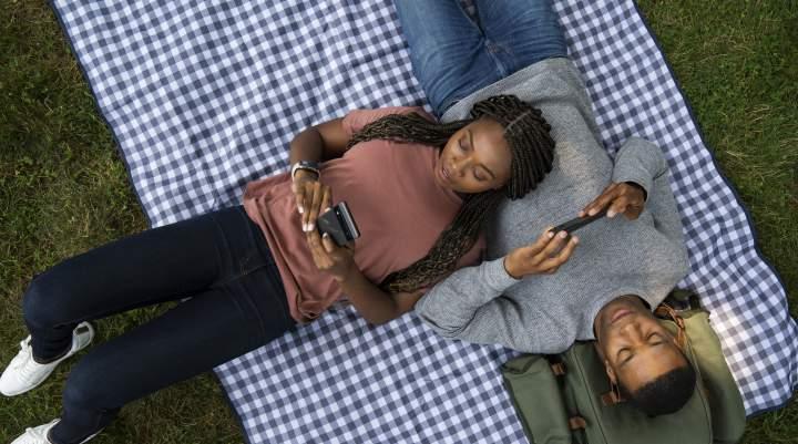 A man and a woman lounge on a picnic blanket playing mobile games.